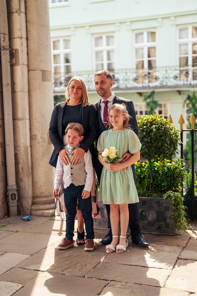 Cute family celebrating outdoors, kids dressed for wedding, smiling with flowers, sunny day, elegant clothing, urban backdrop, happy moments.