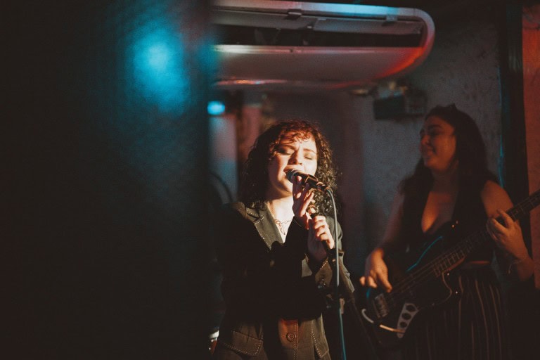 Female singer performing live music in a dimly lit venue with a guitarist in the background.