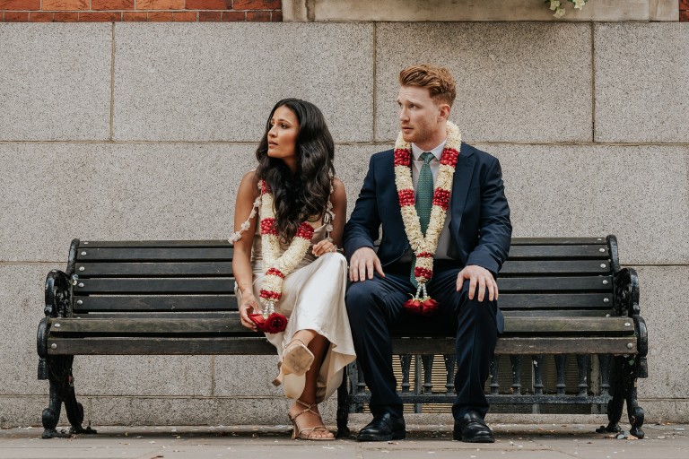 Beautiful diverse couple sitting on a park bench after wedding, wearing floral garlands and elegant attire.