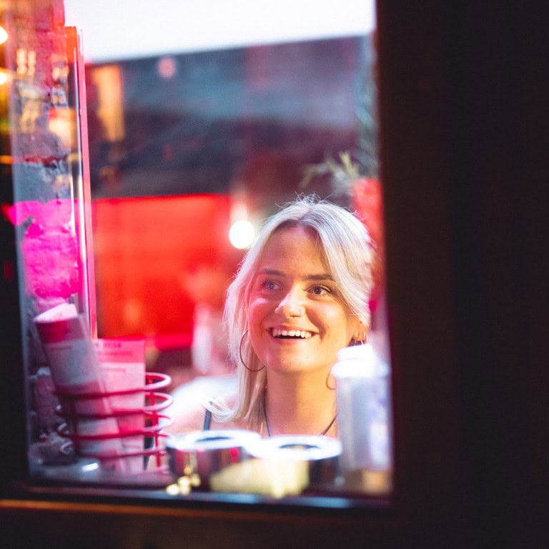 Bright young woman looking through illuminated shop window at night, showcasing nightlife and retail shopping.