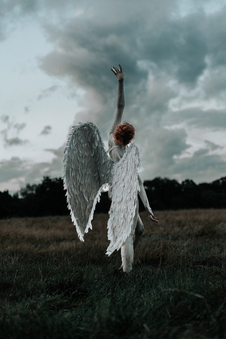 Wings and red hair angel woman standing on grassy field under cloudy sky.