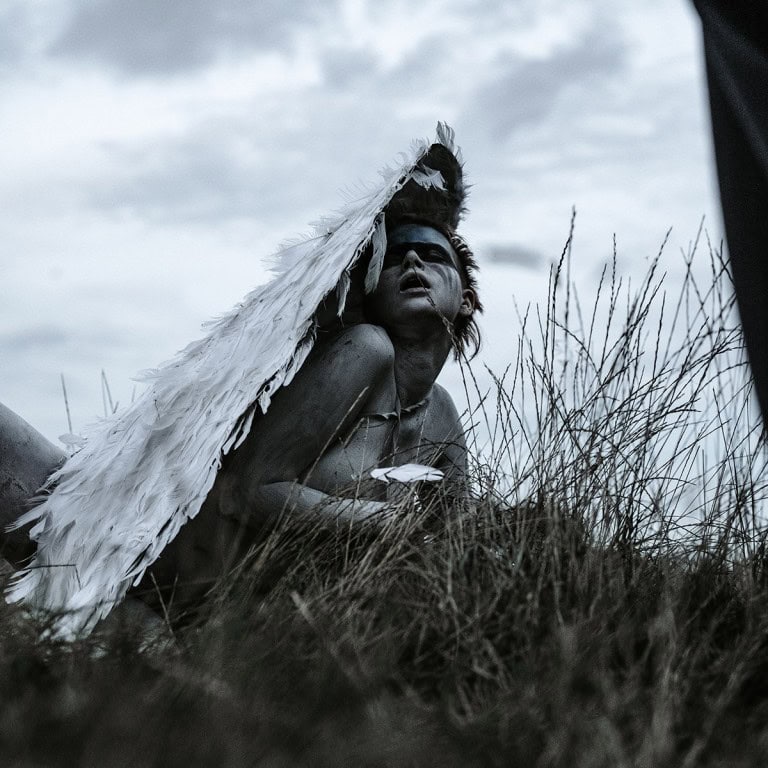 Wounded person lying in dry grass wearing a feathered headdress, black and white photo.