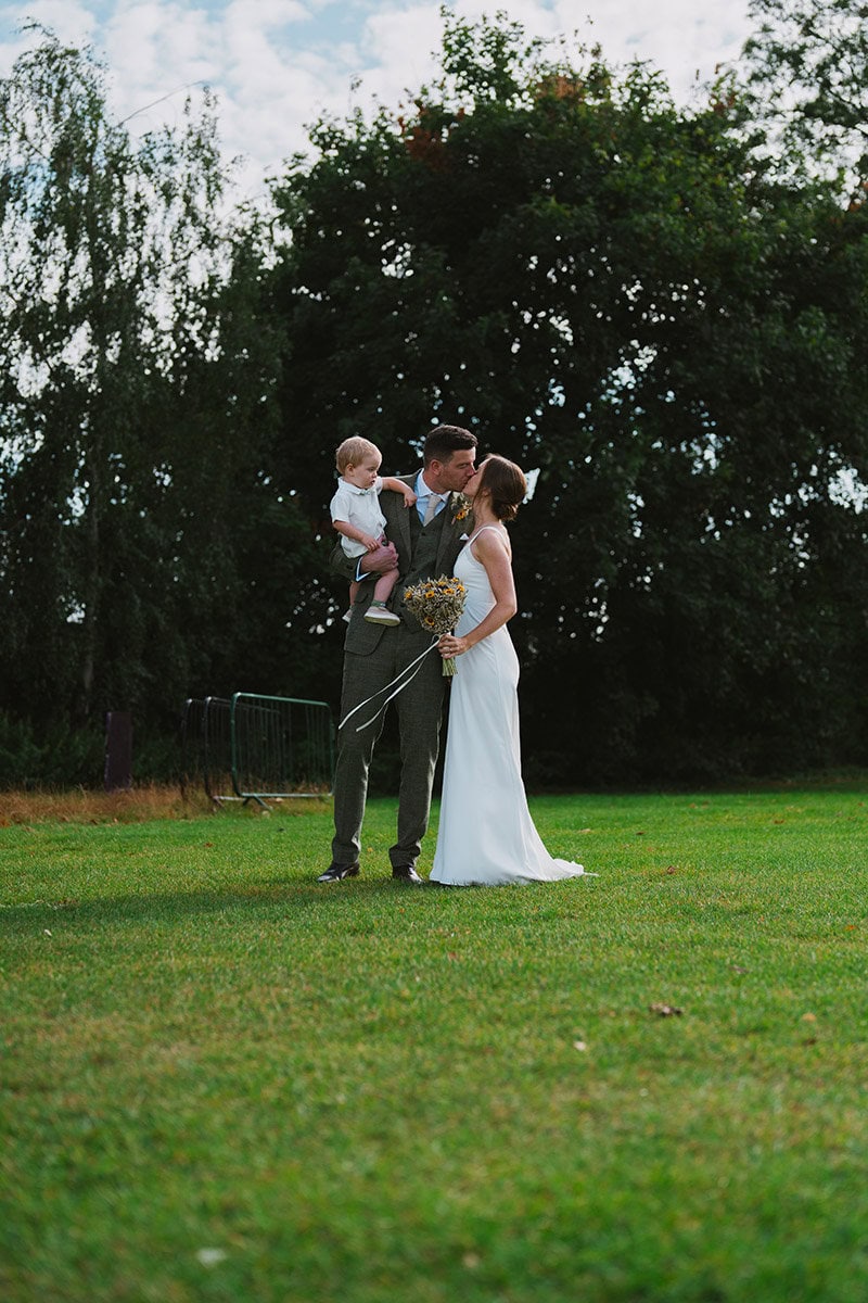 Happy couple kissing during outdoor wedding ceremony with child on bride's arm in lush green park.
