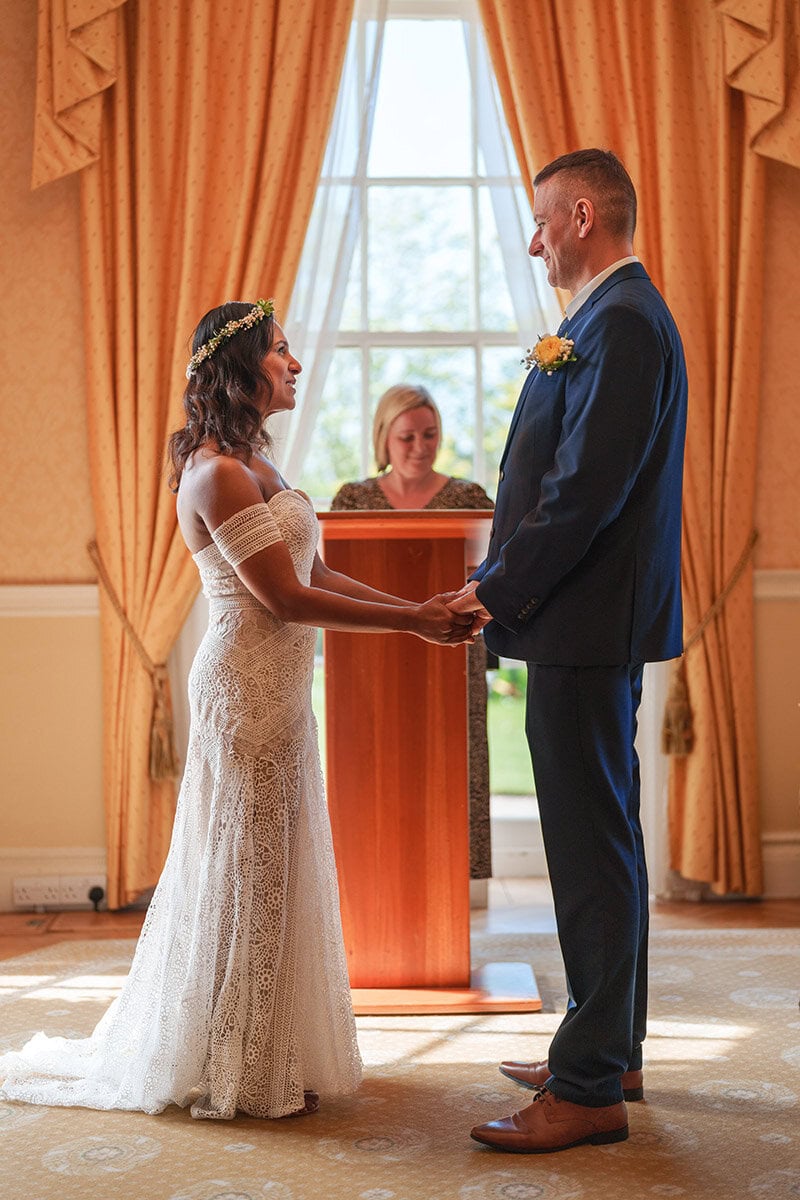 LemonLens wedding ceremony, bride and groom holding hands, intimate wedding moment, indoor setting with elegant drapes.