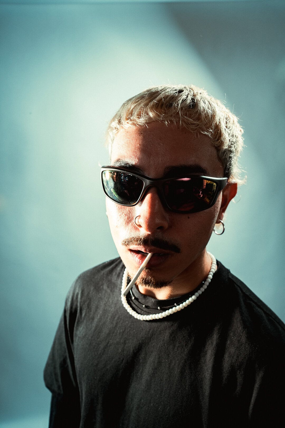 Stylish young man with blonde hair wearing black sunglasses and necklaces, holding a cigarette in his mouth.