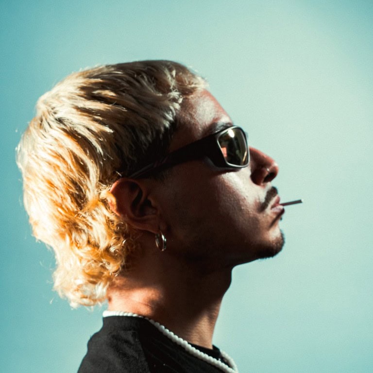 Stylish man with blonde hair, sunglasses, and a cigarette against pastel blue background.
