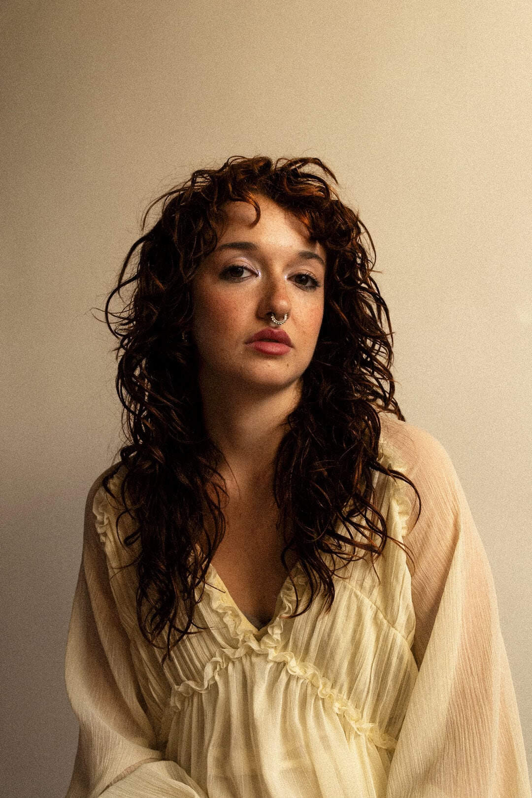 Vibrant portrait of a young woman with curly red hair and septum piercing in a beige blouse, elegant and stylish.