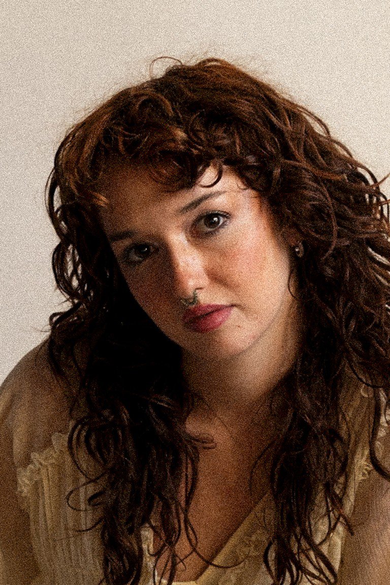 Vibrant woman with curly auburn hair and natural makeup, smiling softly, photographed against a neutral background.