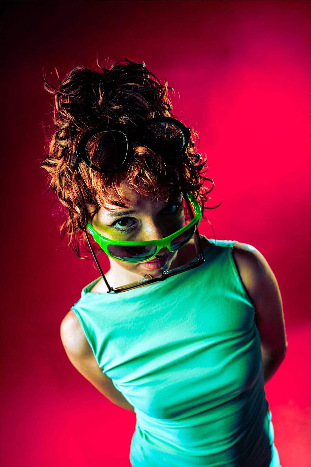 Vibrant woman with curly hair wearing colourful sunglasses against pink background.