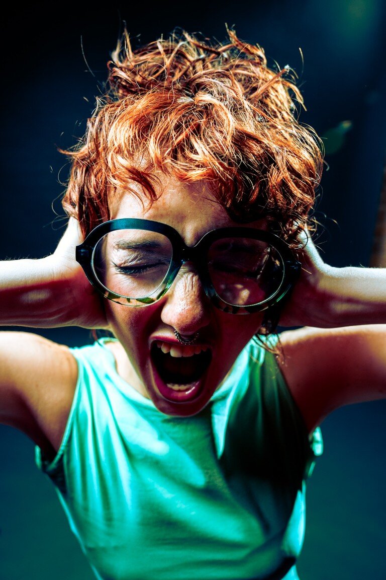 Bright red-haired woman with large glasses screaming with hands behind head, vibrant emotion, expressive portrait.