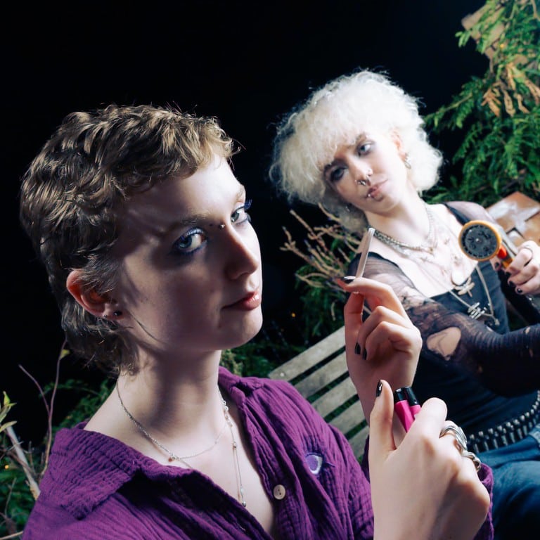 Close-up portrait of two young people with alternative styles, sitting outdoors at night with greenery in the background.