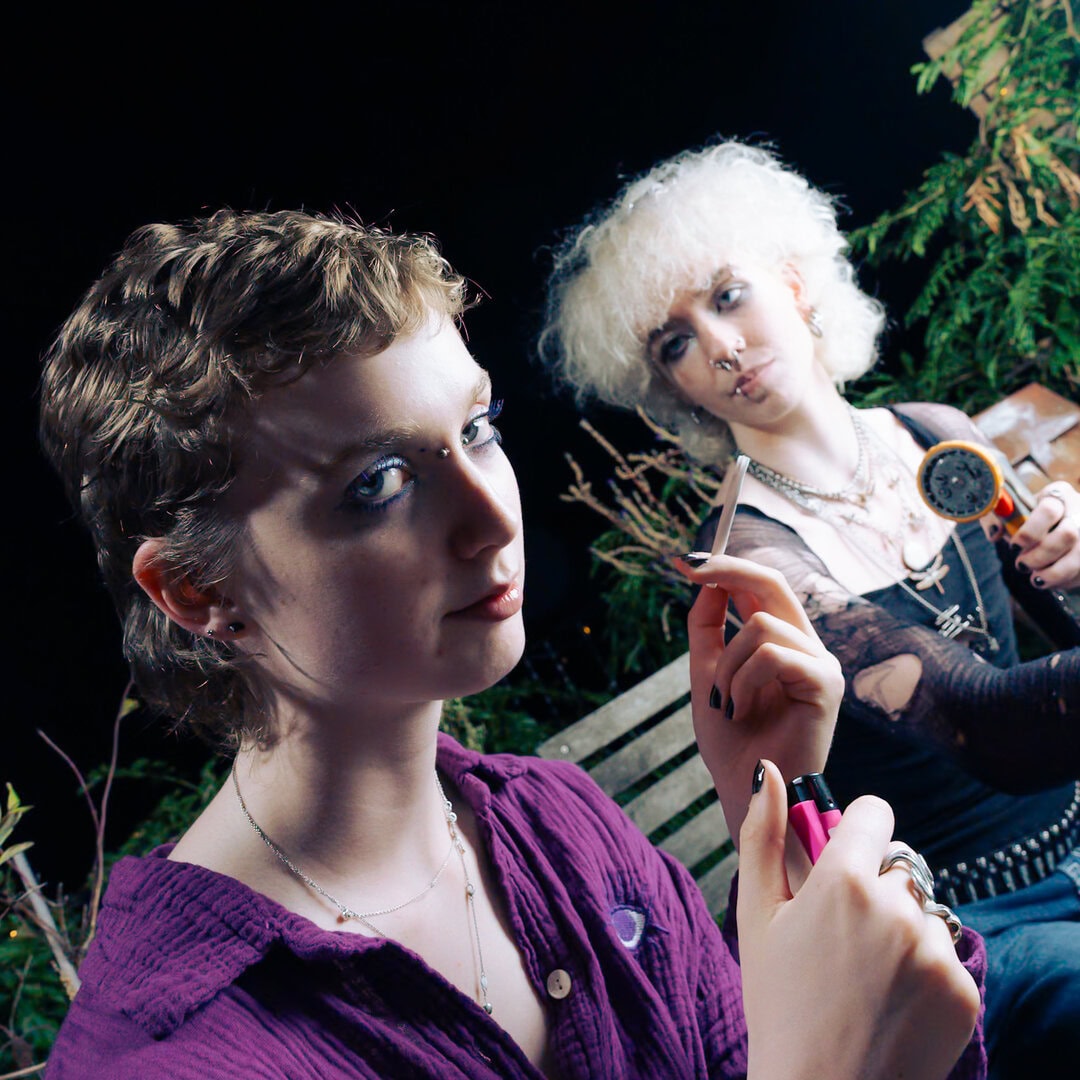 Close-up portrait of two young people with alternative styles, sitting outdoors at night with greenery in the background.