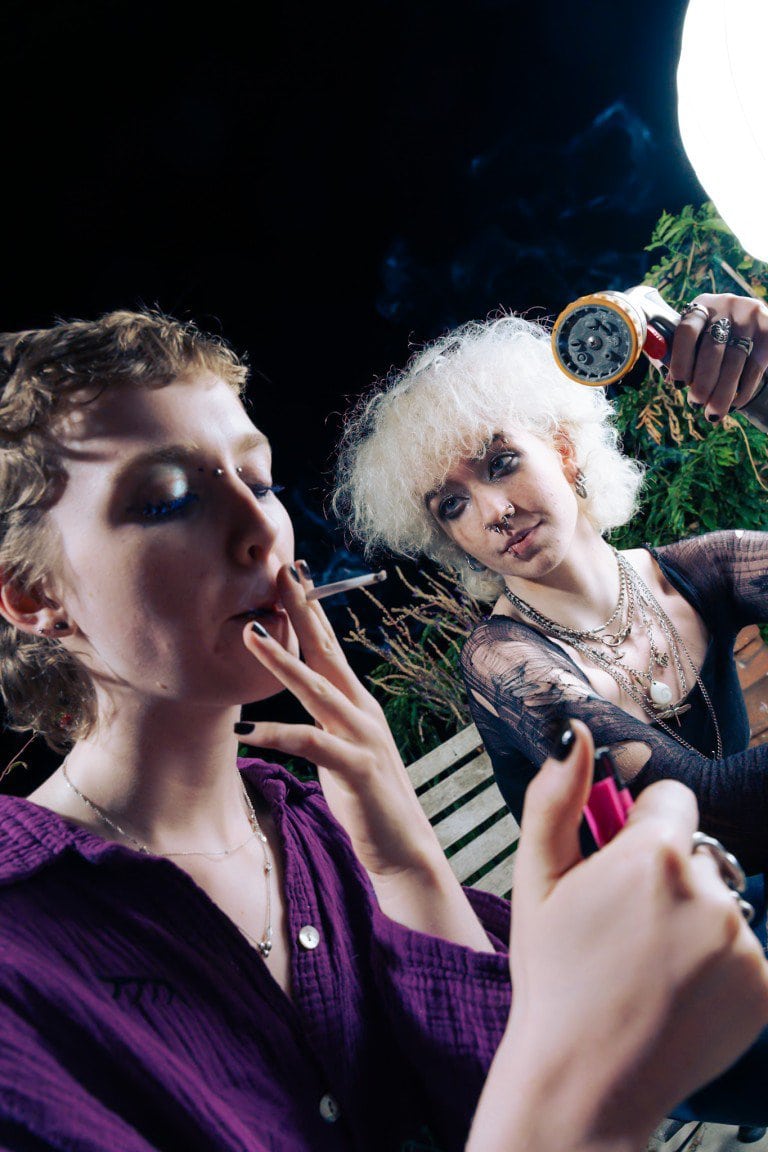 Cigarette smoking, alternative lifestyle, goth fashion, edgy jewellery, grunge style, night-time outdoor setting.