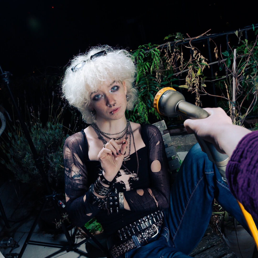 person with peroxide blonde curly hair wearing black distressed top and jewelry sitting outside at night, holding a heat gun, surrounded by plants and wooden benches—moody, alternative style.