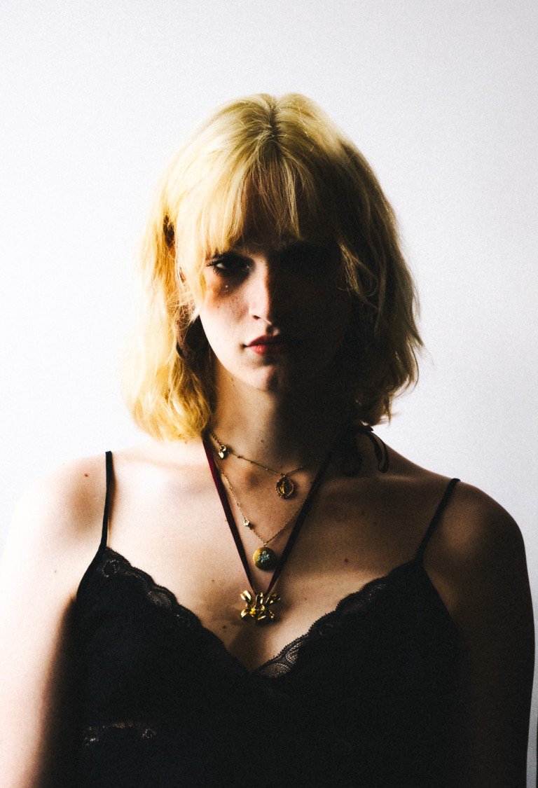 Gothic woman with blonde hair wearing black lace top and layered necklaces.