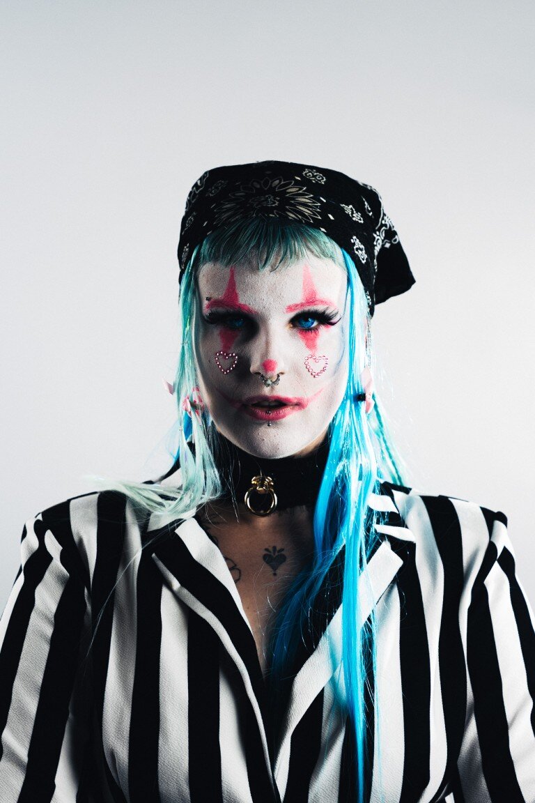Glittery clown makeup with blue hair and piercings, punk fashion, black and white striped jacket, bold alternative style.