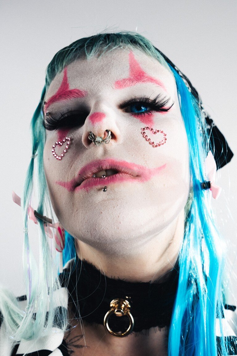 Bright punk-inspired portrait of a person with colourful blue and green hair, vibrant makeup, and extensively piercings.