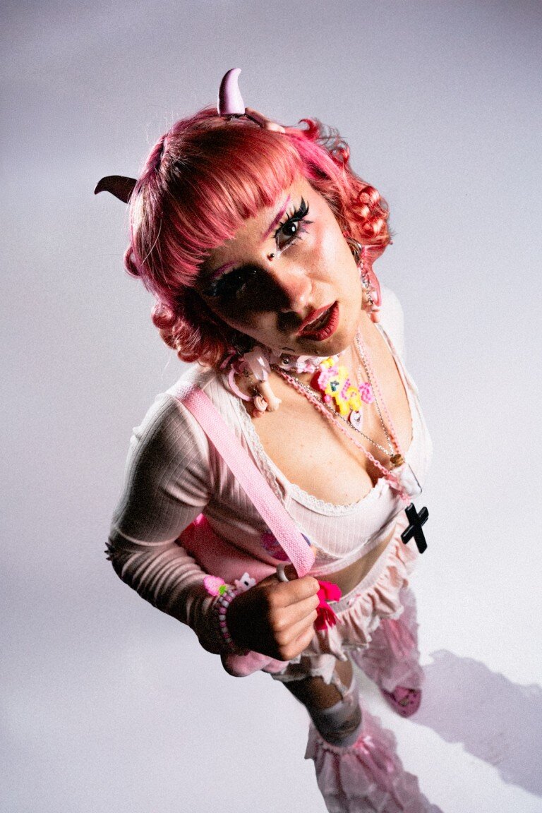 Bright pink-haired woman with unicorn horn accessories and colourful jewellery, dressed in pastel and Gothic Lolita style.