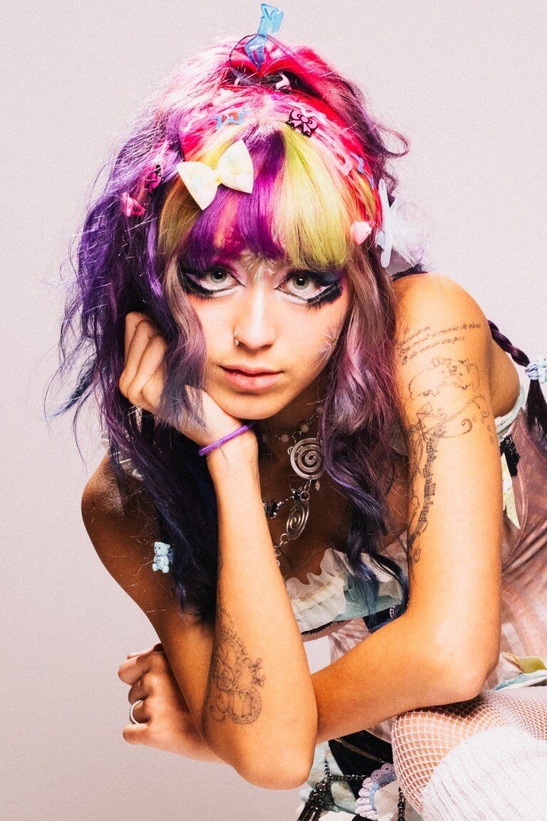 Vibrant rainbow-haired young woman with tattoos, colourful hair accessories, and artistic makeup.