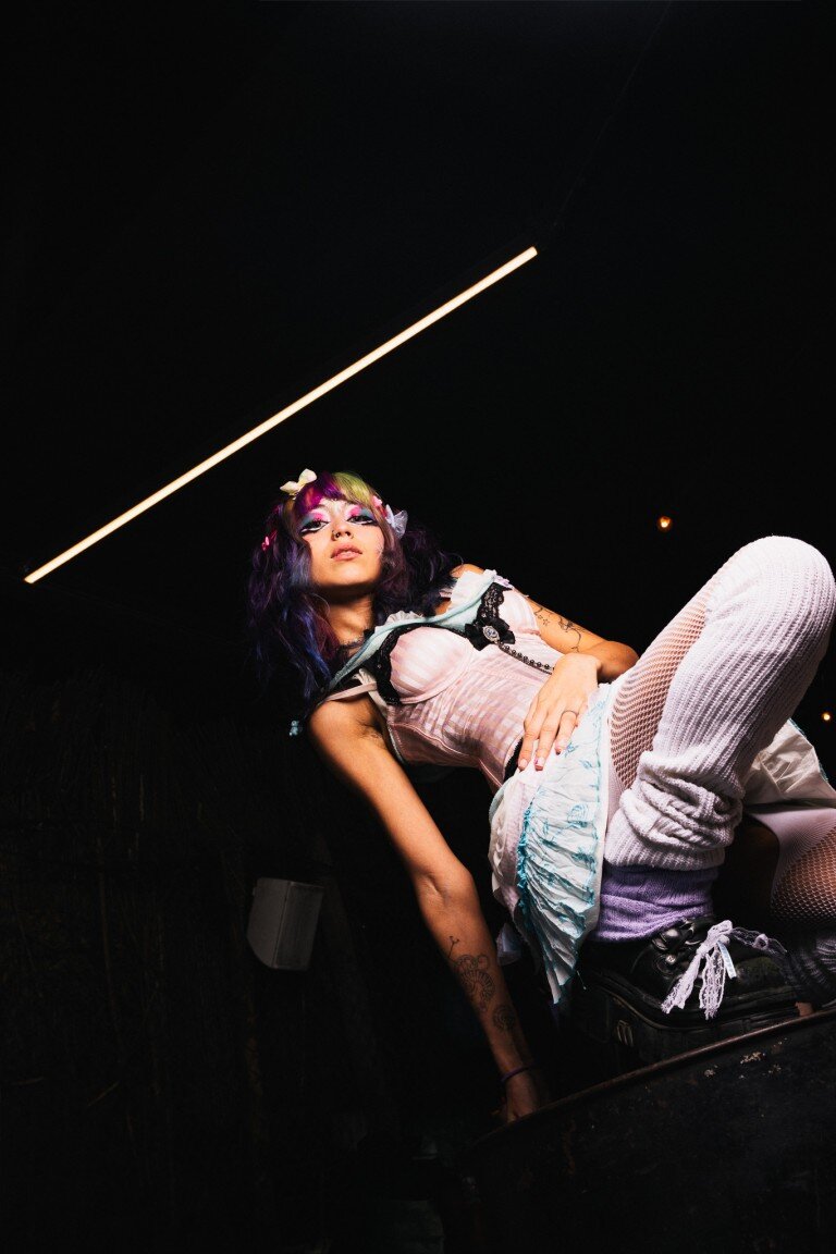 Colourful edgy woman with tattoo, neon makeup, black hair with purple and green tips, in dark room with modern lighting.