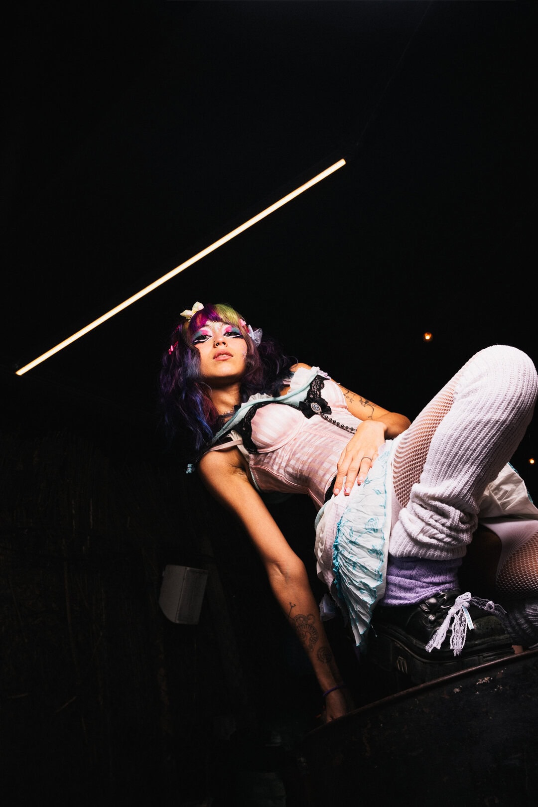 Colourful edgy woman with tattoo, neon makeup, black hair with purple and green tips, in dark room with modern lighting.