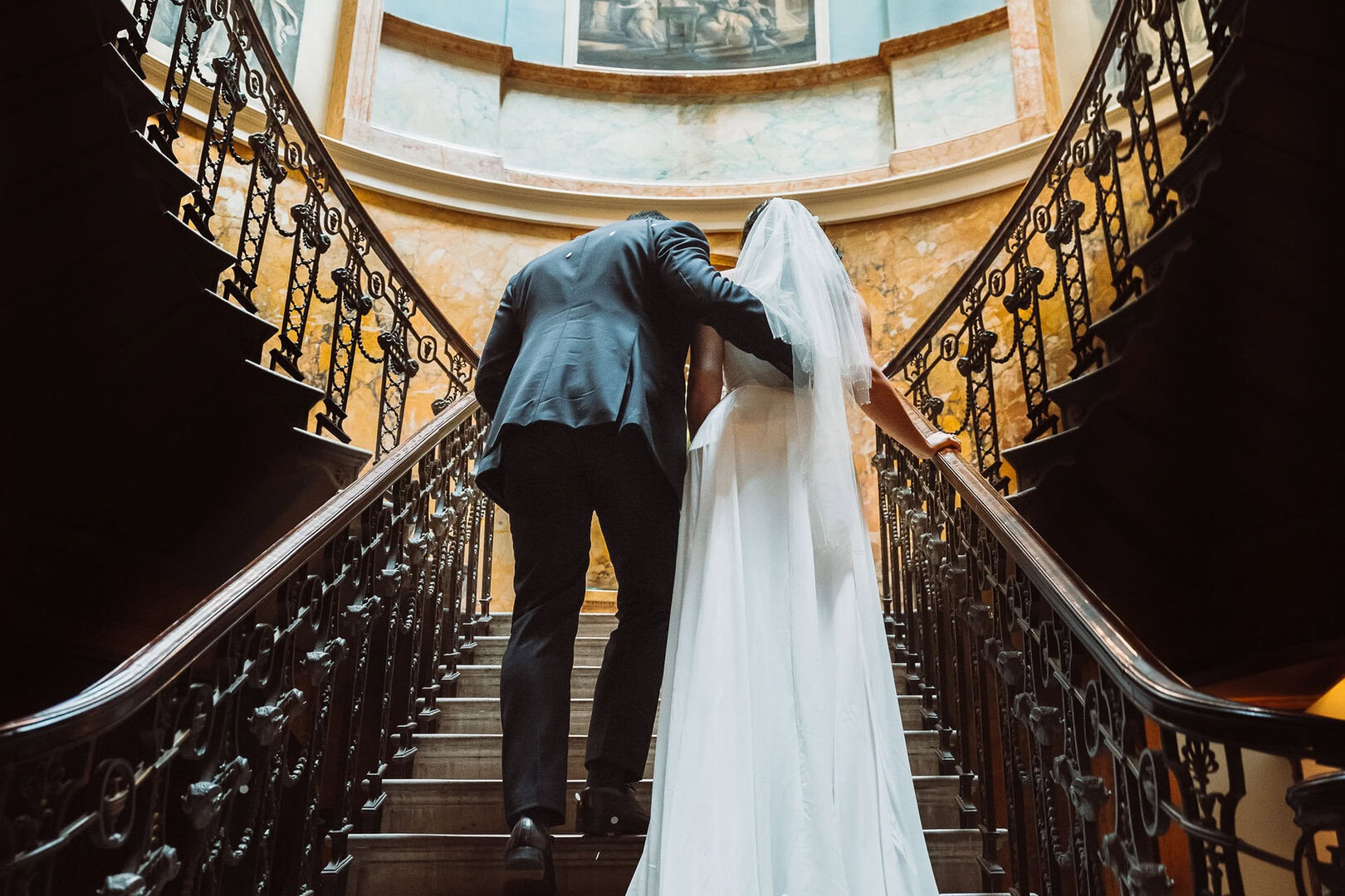 Elegant wedding couple ascending ornate staircase in historic venue, bride in white dress and veil, groom in suit, romantic moment.
