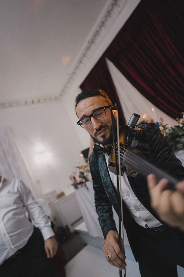 Elegant man playing violin at wedding reception, dressed in tuxedo, joyful atmosphere, wedding decor in background.