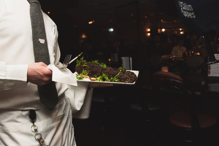 Plate of blackened fish garnished with fresh herbs served by a waiter in a dimly lit restaurant.