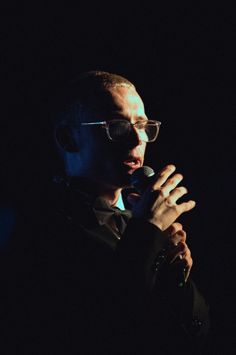 Professional singer performing with microphone in dark setting, wearing glasses, bow tie and suit.