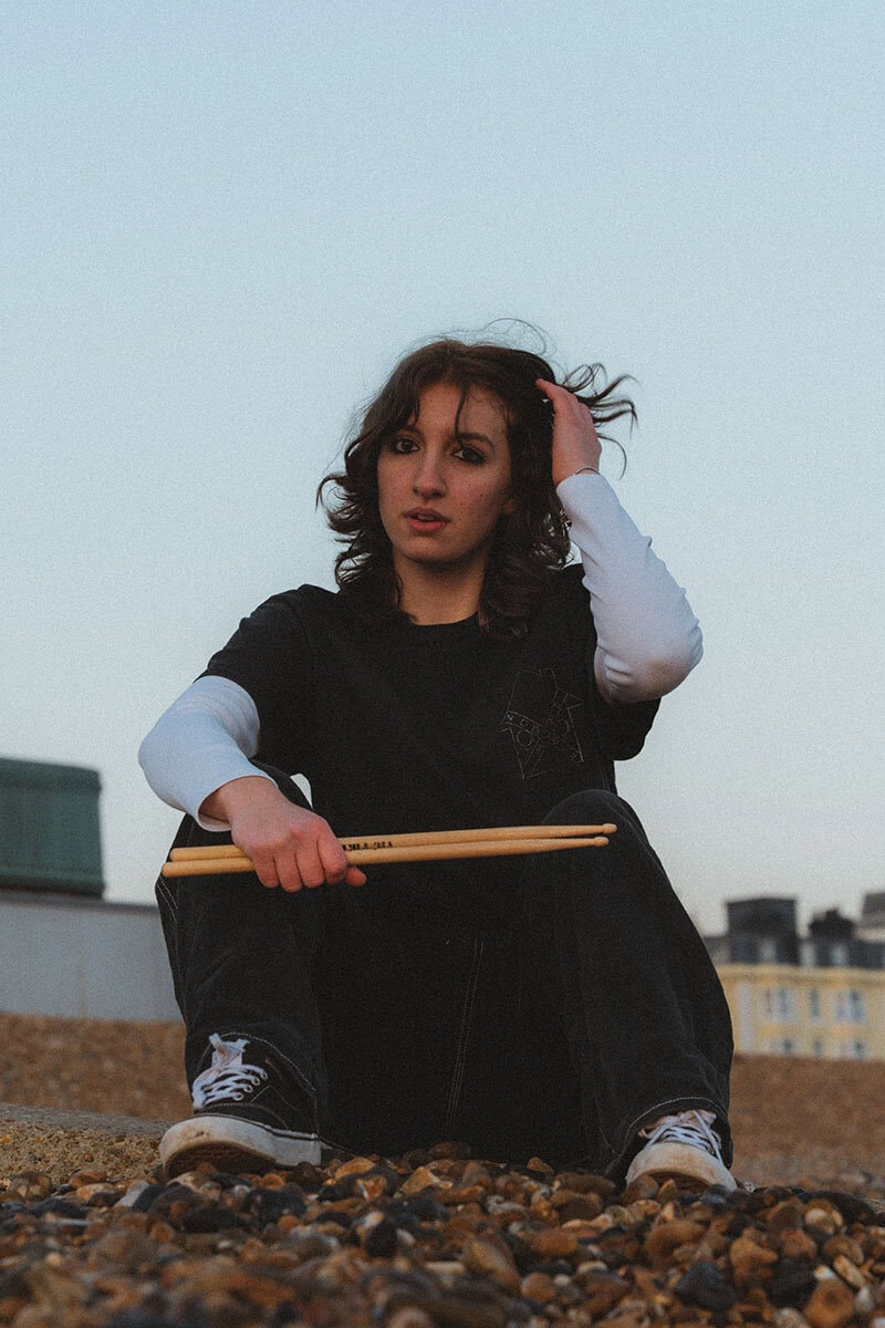 Drummer woman sitting on rocky beach with drumsticks in hand.