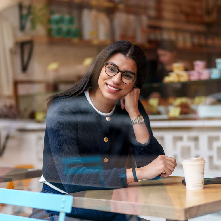 Woman smiling sitting at a cafe table; casual style, glasses, coffee cup, warm atmosphere.