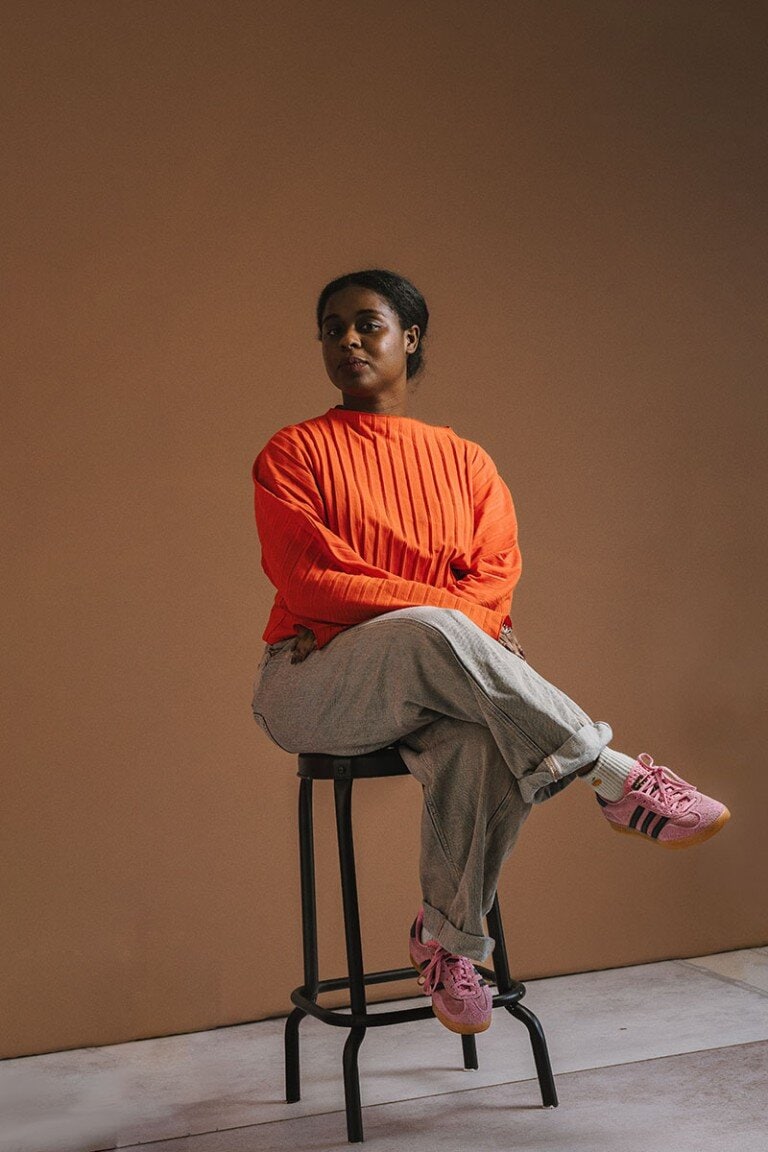 Bright orange sweatshirt and grey joggers worn by woman sitting on stool against brown background.