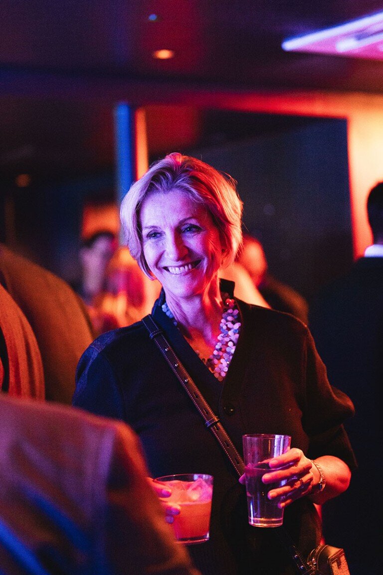 Bright smiling woman at a lively party holding a drink in a colourful, vibrant nightclub scene.