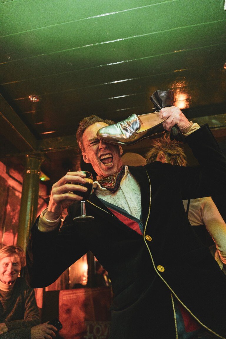 Vibrant party scene with a man in flamboyant attire, holding a glass of red wine and a shoe, celebrating joyfully at a lively event.