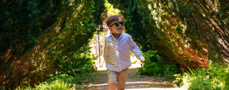 Bright young boy wearing sunglasses walking through a green outdoor tunnel of foliage.