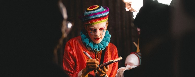 An artist with colorful clown makeup and vibrant attire, writing in a notebook at LemonLens.