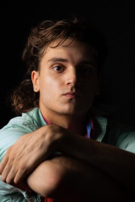 Young man with wavy brown hair and clear skin, resting with a contemplative expression against dark background.