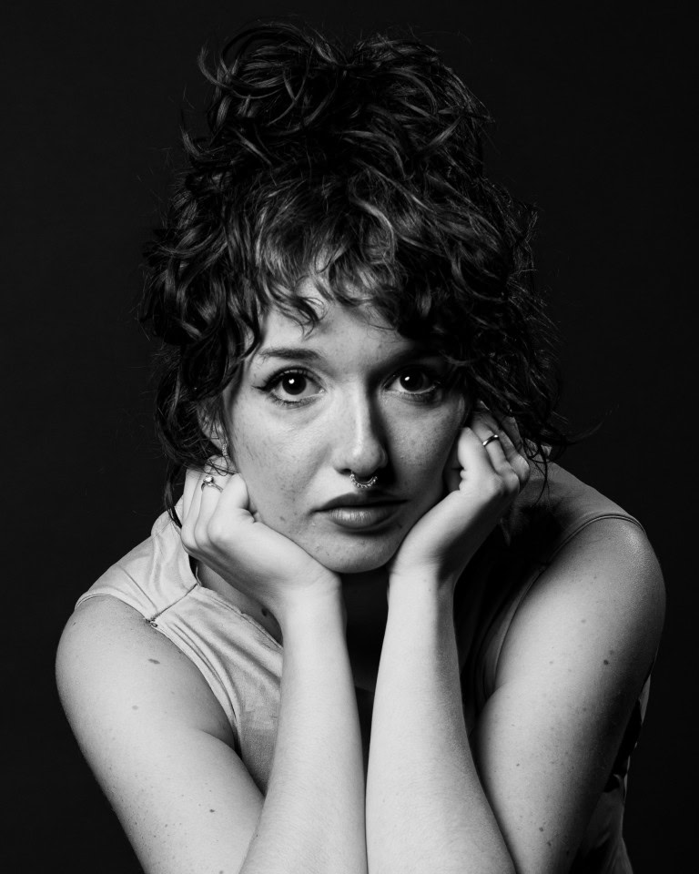 Close-up black and white portrait of a young woman with curly hair, expressive eyes, freckles, and septum piercing.
