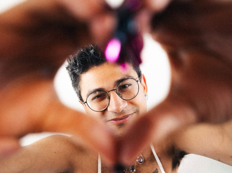 Close-up of a young person with glasses, looking through hands made into a heart shape, with soft lighting and a blurred background.