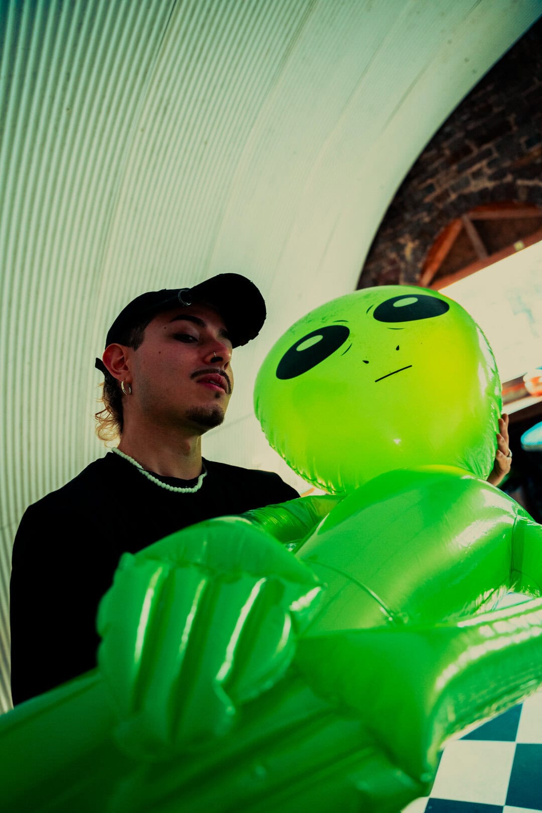 Bright green inflatable alien figure with a person holding it, in an indoor setting with a curved ceiling.