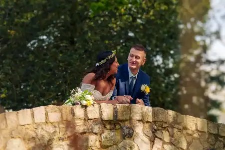 Romantic wedding couple leaning on stone wall in outdoor greenery scene.