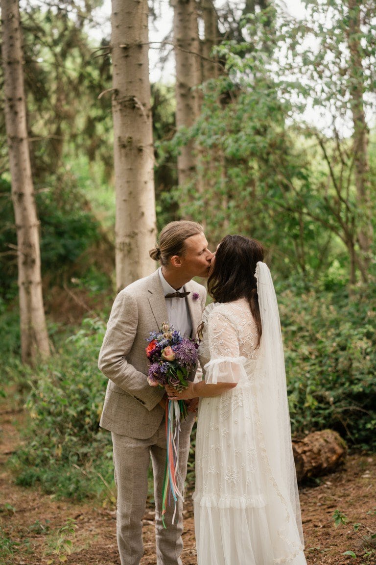 Romantic wedding couple sharing a kiss in a forest setting with trees and lush greenery.