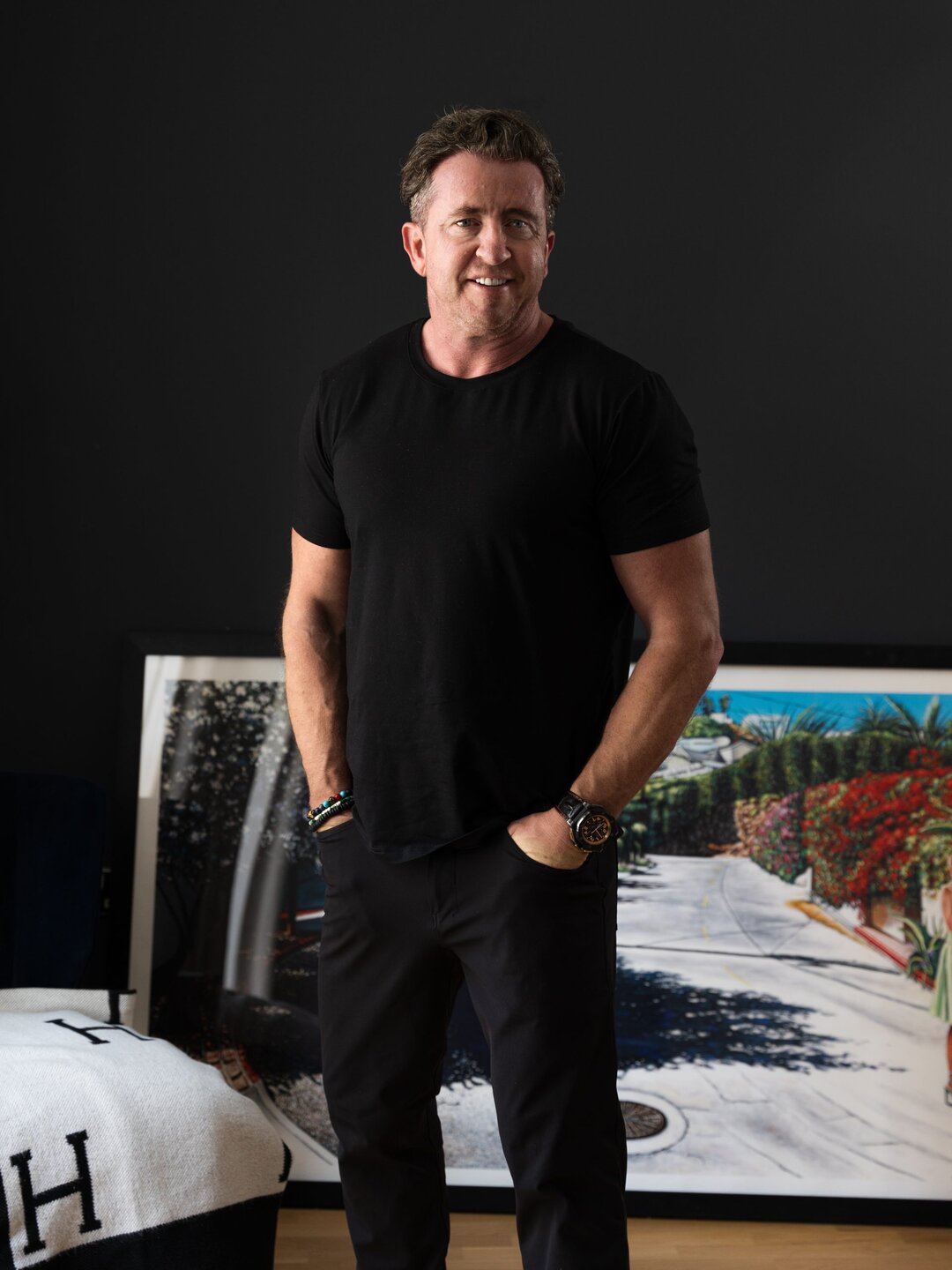 Portrait of a smiling man wearing a black t-shirt and accessories, standing in front of colourful artwork.