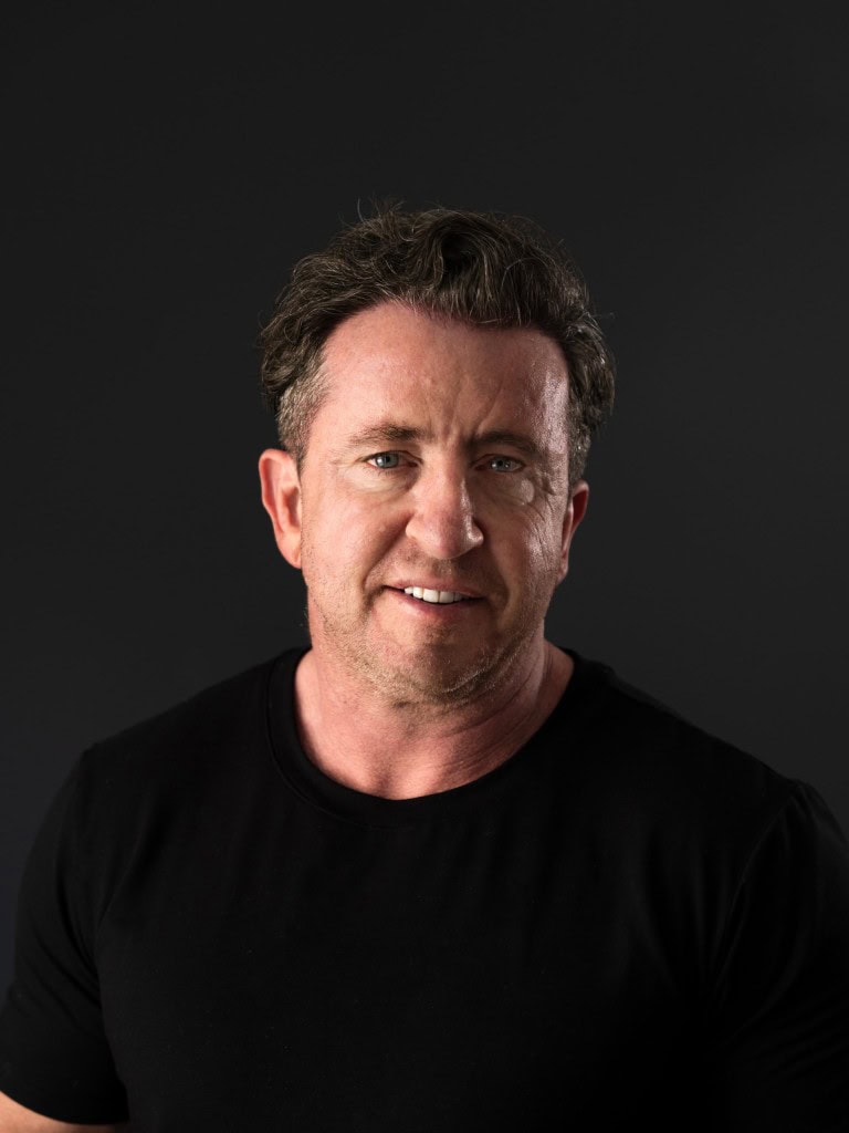 A smiling man with tousled dark hair wearing a black t-shirt against a dark background.