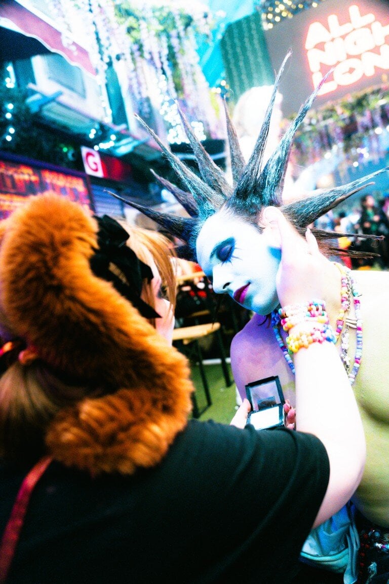 Vibrant nightlife scene with a performer in bold makeup and spiked hairstyle, being photographed at a colourful event.