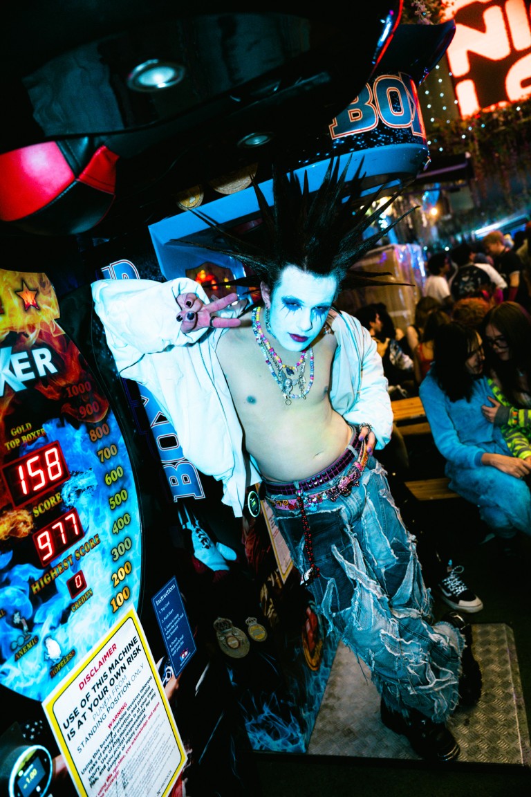 Wild punk performer with spiked hair and blue face paint at an arcade, colourful festival atmosphere.