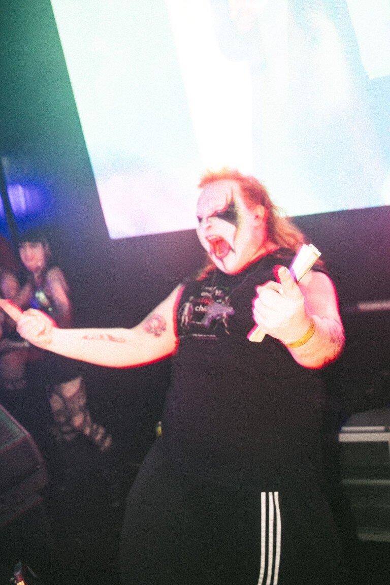 Colorful performer with face paint at a music event, holding drumsticks.