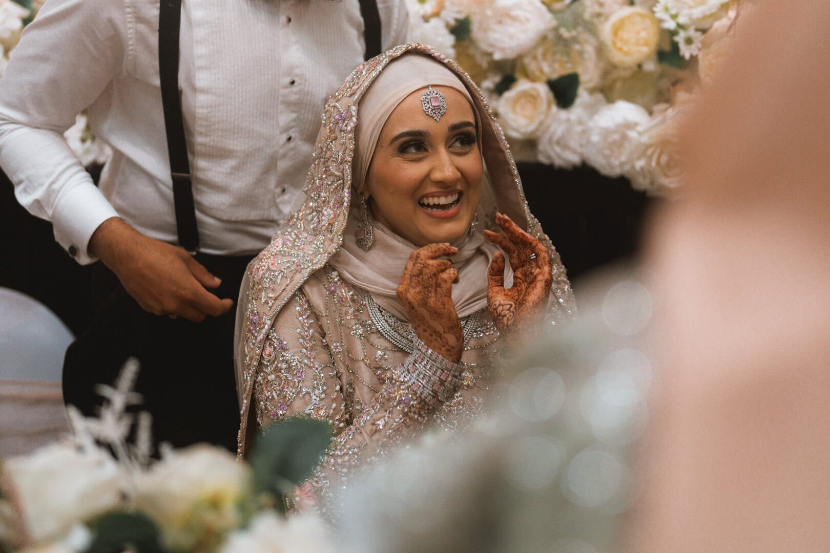 Elegant woman in floral wedding attire with intricate henna designs, smiling amid a backdrop of white roses.