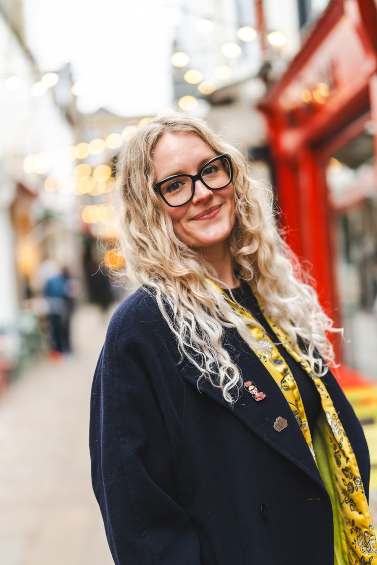 Bright-eyed individual with curly blonde hair and black glasses smiles warmly on a lively street filled with festive lights and colourful buildings.