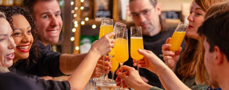 Chefs and diners clinking glasses with bright orange drinks in a lively restaurant setting.